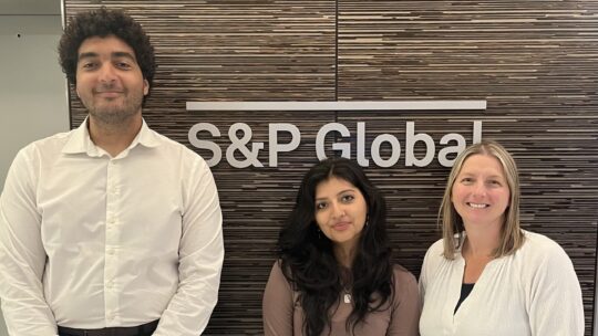 Three people stand in front of the S&P Global sign